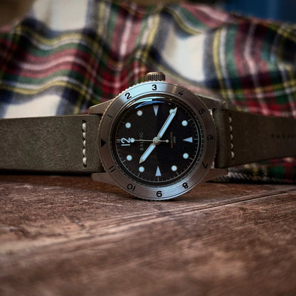 Watch with a grey leather strap on a wooden surface, with a plaid shirt in the background.