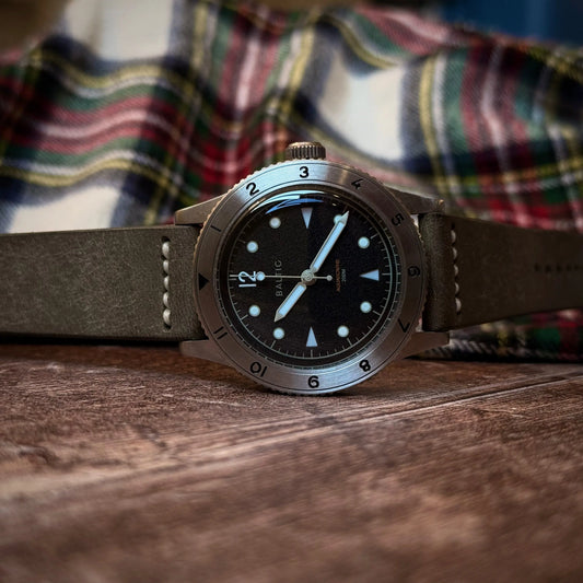 Watch with a grey leather strap on a wooden surface, with a plaid shirt in the background.